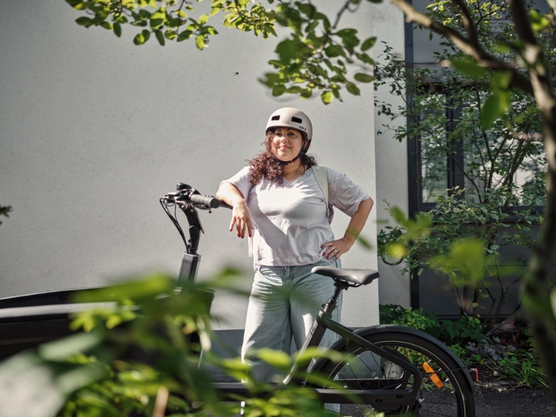 Frau mit Locken lehnt am JobRad, im Vordergrund Pflanzen Frau mit Locken lehnt am JobRad, im Vordergrund Pflanzen