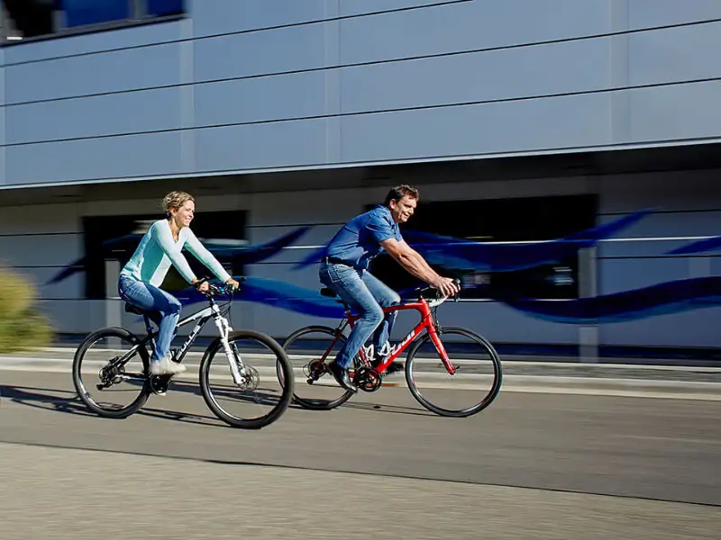 Zwei Menschen, die auf zwei Stadträdern vor einem Büro-Gebäude vorbeifahren Zwei Menschen, die auf zwei Stadträdern vor einem Büro-Gebäude vorbeifahren